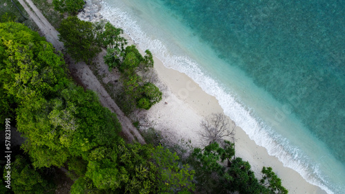 Aerial view of crystal clear turquoise ocean water lined by green forest trees and waves rolling onto white sandy coastline beach on a remote tropical island