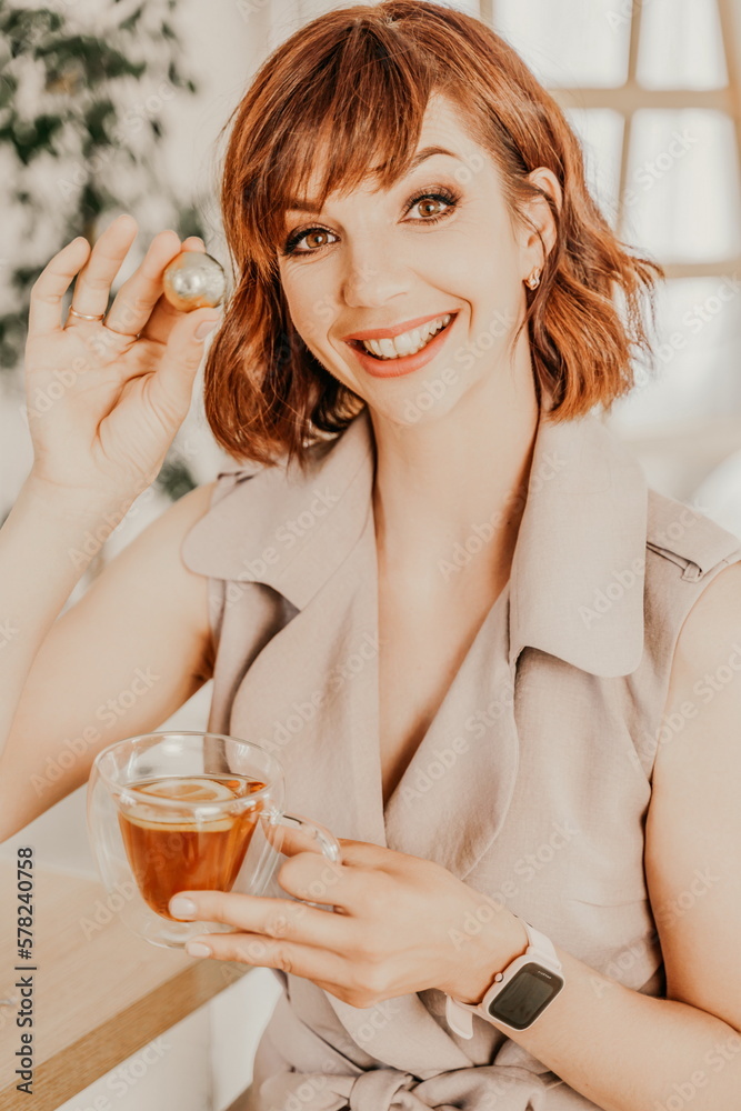 Fototapeta premium Woman drinks tea close-up. Portrait of a brunette in a beige dress with a transparent mug and a candy in her hands.