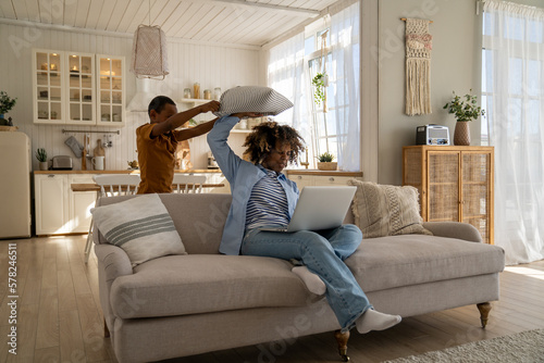 Картината върху платно Exhausted displeased mother sitting on couch at home working on laptop while little child distracts