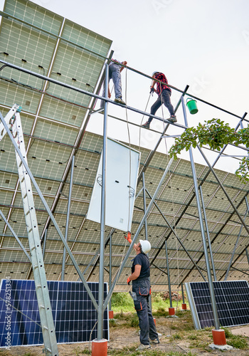 Male solar technicians standing on metal rails and lifting solar module while man helping colleagues. Solar installers team mounting solar panels on metal construction. Concept of renewable energy.