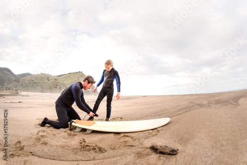 Adult man helps girl fasten surfboard leash at a beach