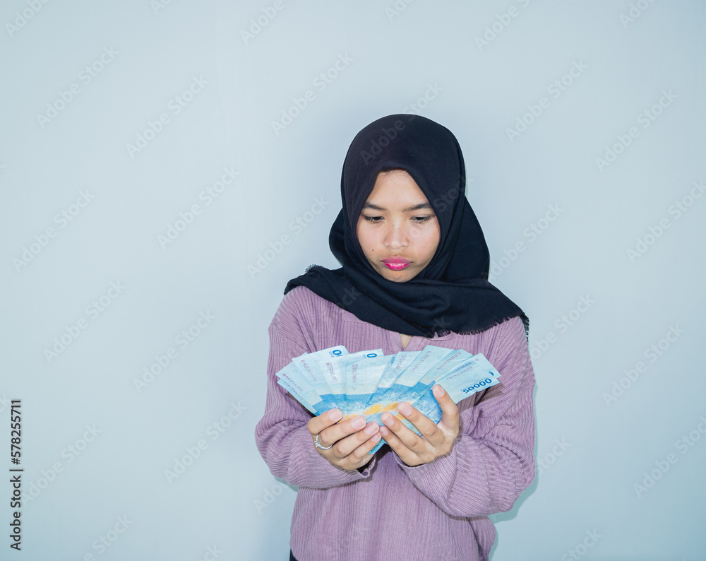Obraz premium Cheerful beautiful Asian woman in a hijab showing Indonesian rupiah banknotes, celebrating luck isolated over white background. People religious lifestyle concept. ramadan concept. holiday allowance