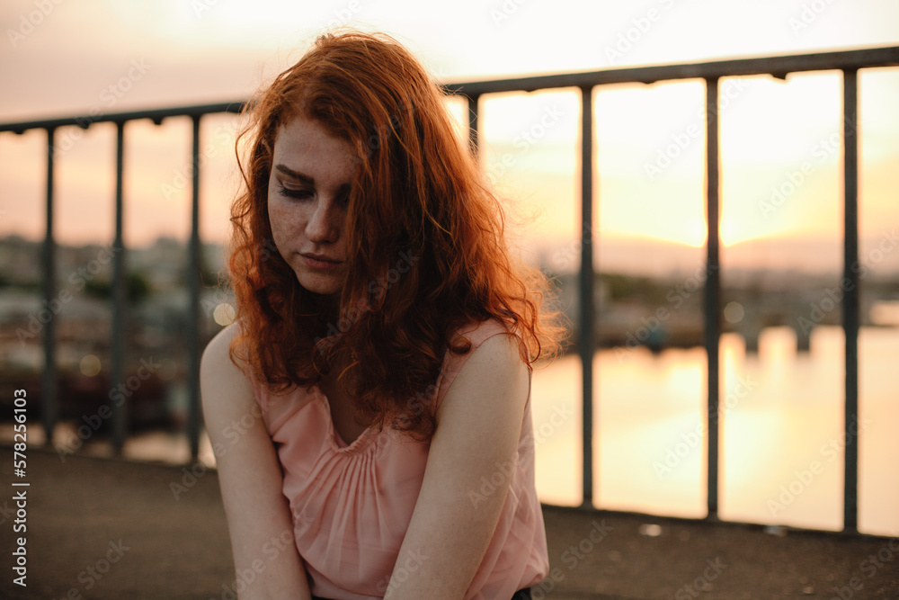 Portrait of young woman with red hair sitting on bridge at sunset