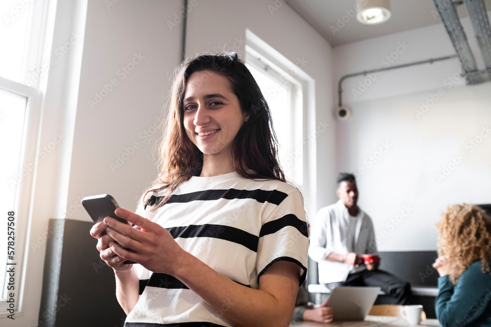 Happy businesswoman holding smart phone with colleagues in background Happy businesswoman holding smart phone with colleagues in background