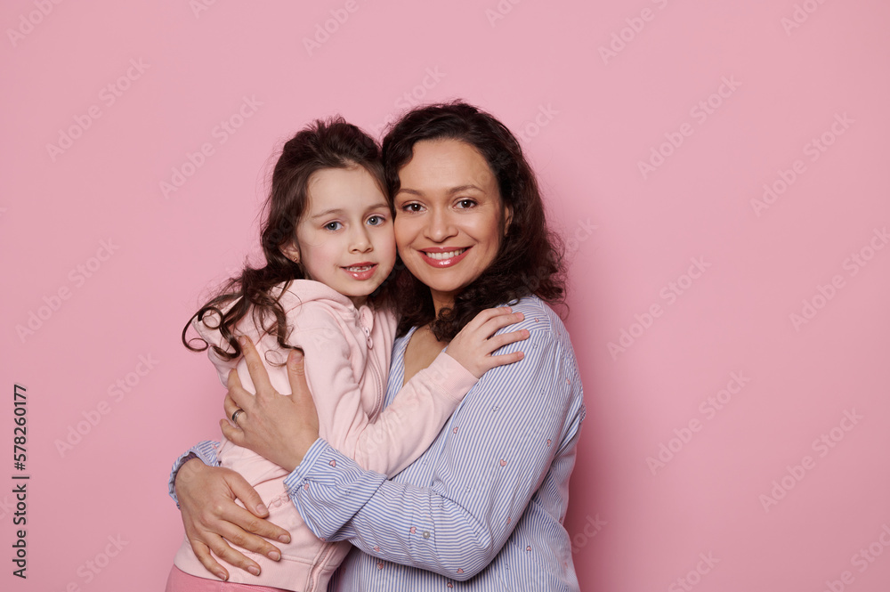 Loving mother hugging her little daughter, cutely smiling looking at camera, standing together on isolated pink background. Copy space. Happy family relationships. Mother's and Children's Day concept Loving mother hugging her little daughter, cutely smiling looking at camera, standing together on isolated pink background. Copy space. Happy family relationships. Mother's and Children's Day concept