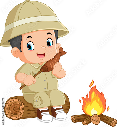 an adventurous boy sits on a fallen tree and is eating grilled fish in front of a bonfire