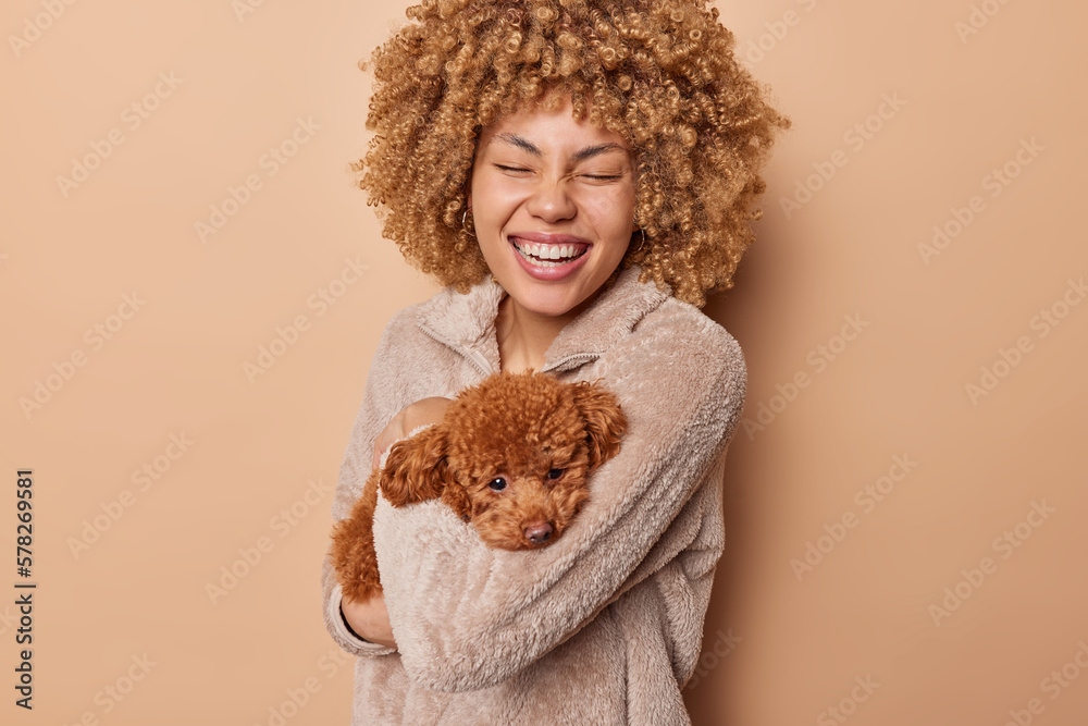 Portrait of cheerful curly haired young woman laughs happily expresses positive emotions has fun holds sad poodle puppy on hands isolated over brown background. People and favorite pets concept
