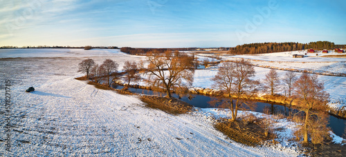 Fototapeta Naklejka Na Ścianę i Meble -  River flowing in snowy winter fields in wilderness at sunset. Stream meandering frosty countryside on cold evening. Aerial view of trees on riverbank.
