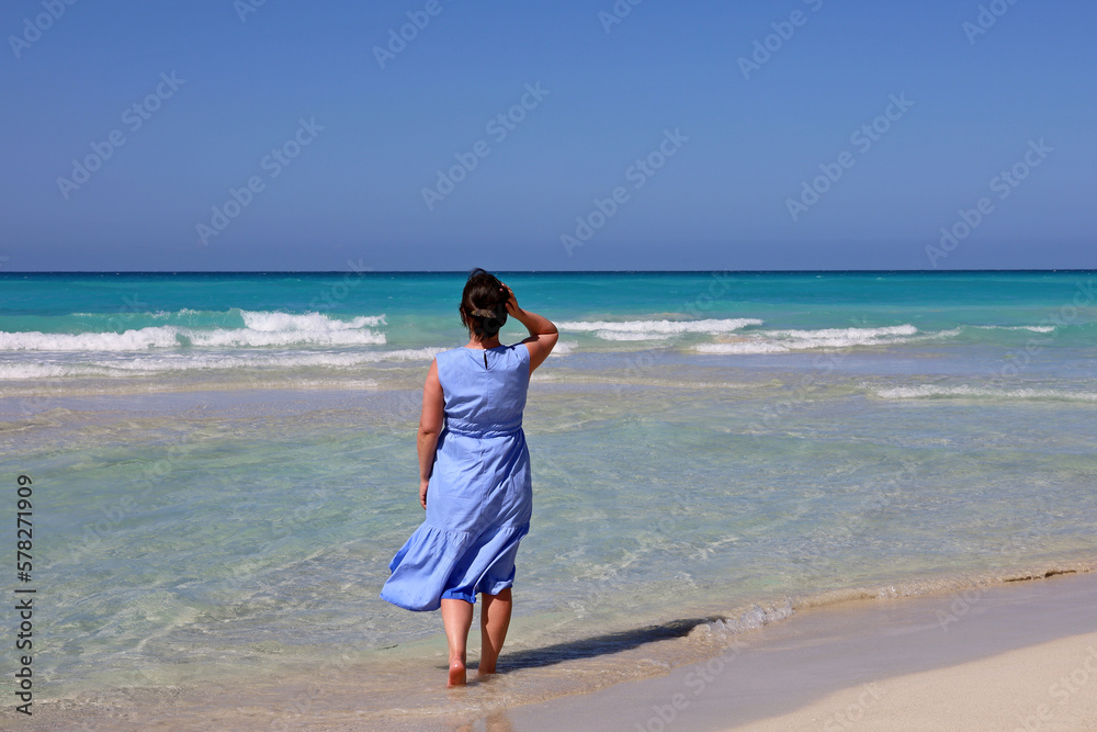 Barefoot woman in blue dress walking by sand against the azure waves. Travel and beach vacation on ocean coast Barefoot woman in blue dress walking by sand against the azure waves. Travel and beach vacation on ocean coast