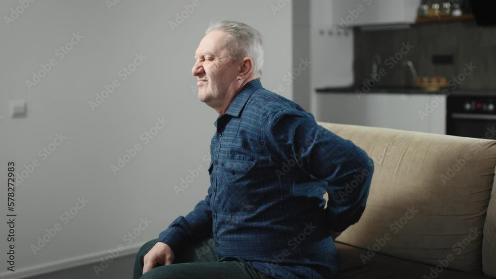 An elderly man sits on the couch at home and has back pain. A man in a shirt at home, suffering from pain An elderly man sits on the couch at home and has back pain. A man in a shirt at home, suffering from pain