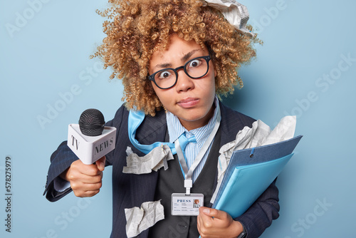 Shocked woman with curly hair stares at camera wears spectacles and formal outfit holds folders and microphone prepares for taking interview or giving speech during live stream. Mass media concept