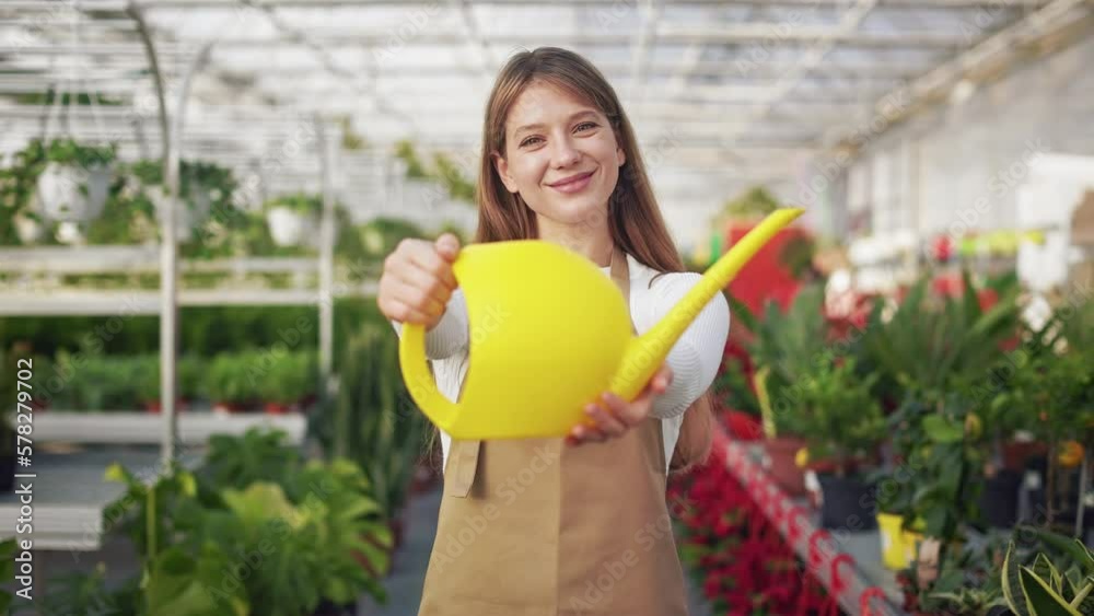 Front view of attractive, young florist enjoying work in greenhouse, orangery. Cheerful, positive lady holding watering pot, showing, smiling. Concept of happiness and joy. Front view of attractive, young florist enjoying work in greenhouse, orangery. Cheerful, positive lady holding watering pot, showing, smiling. Concept of happiness and joy.