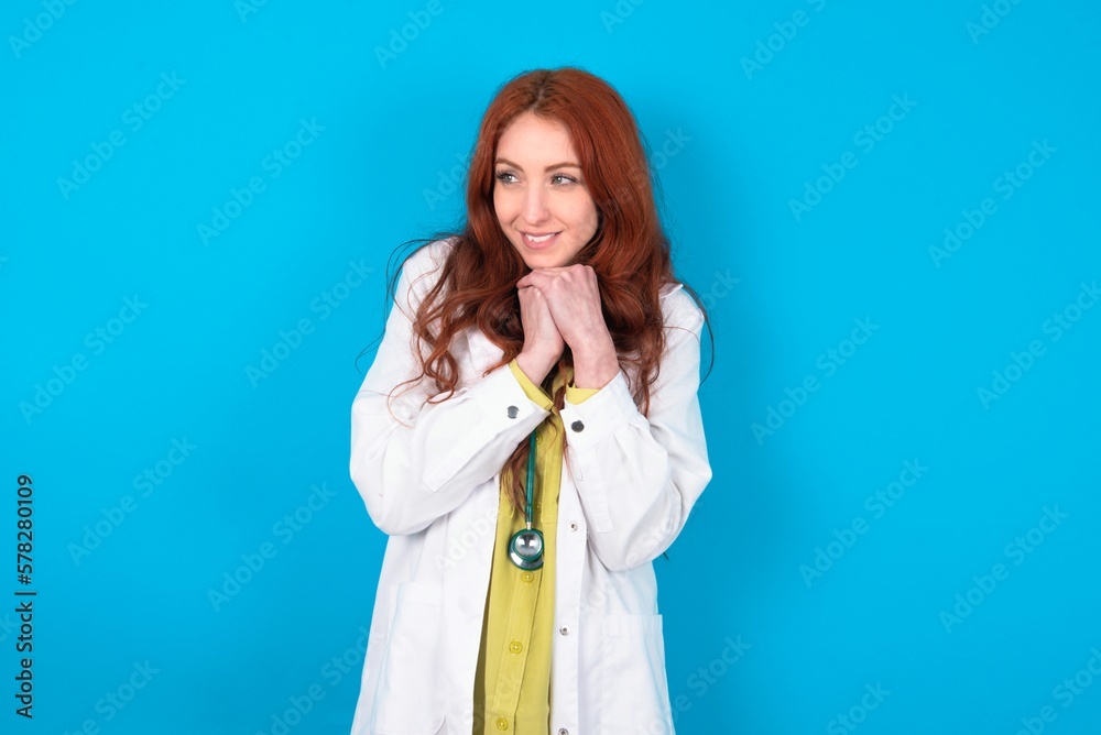 Happy young doctor woman wearing medical uniform over blue background anticipates something awesome happen, looks happily aside, keeps hands together near face, has glad expression. Happy young doctor woman wearing medical uniform over blue background anticipates something awesome happen, looks happily aside, keeps hands together near face, has glad expression.