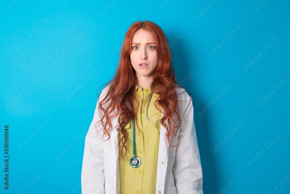 Shocked young doctor woman wearing medical uniform over blue background stares bugged eyes keeps mouth opened has surprised expression. Omg concept Shocked young doctor woman wearing medical uniform over blue background stares bugged eyes keeps mouth opened has surprised expression. Omg concept