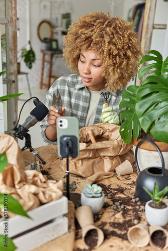 Famous curly female blogger gives gardening classes online shows gardening tools records tutorial video uses microphone and smartphone surrounded by pots with soil poses against home interior