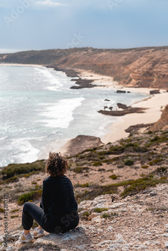 Women with short hair and black tshirt sitting in the rocks closed to the beach and looking to the sea in a magical landscape