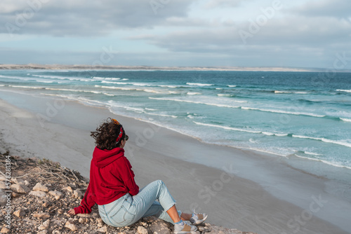 Women with short hair and red hoodie sitting in the rocks closed to the beach and looking to the sea