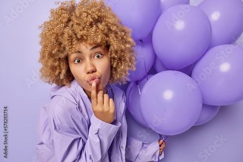 Portrait of good looking surprised woman keeps lips folded stares impressed dressed in anorak holds bunch of inflated balloons celebrates special occasion isolated over purple background. Monochrome
