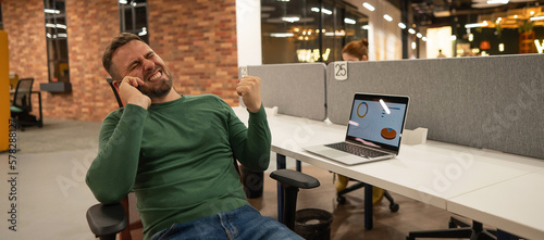Caucasian bearded man talking on a smartphone while sitting at his desk in a modern coworking space.