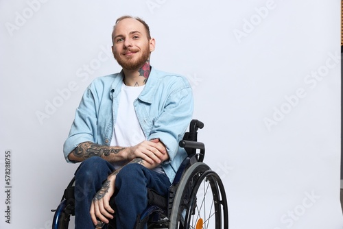 Obraz na plátne A man in a wheelchair smile, copy space, with tattoos on his arms sits on a gray