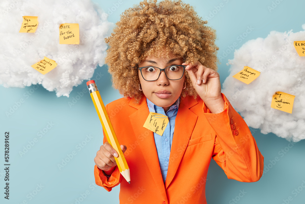 Shocked woman keeps hand on rim of spectacles stares impressed holds big wooden pencil wears orange jacket cannot believe own eyes isolated over blue background white clouds overhead sticky notes