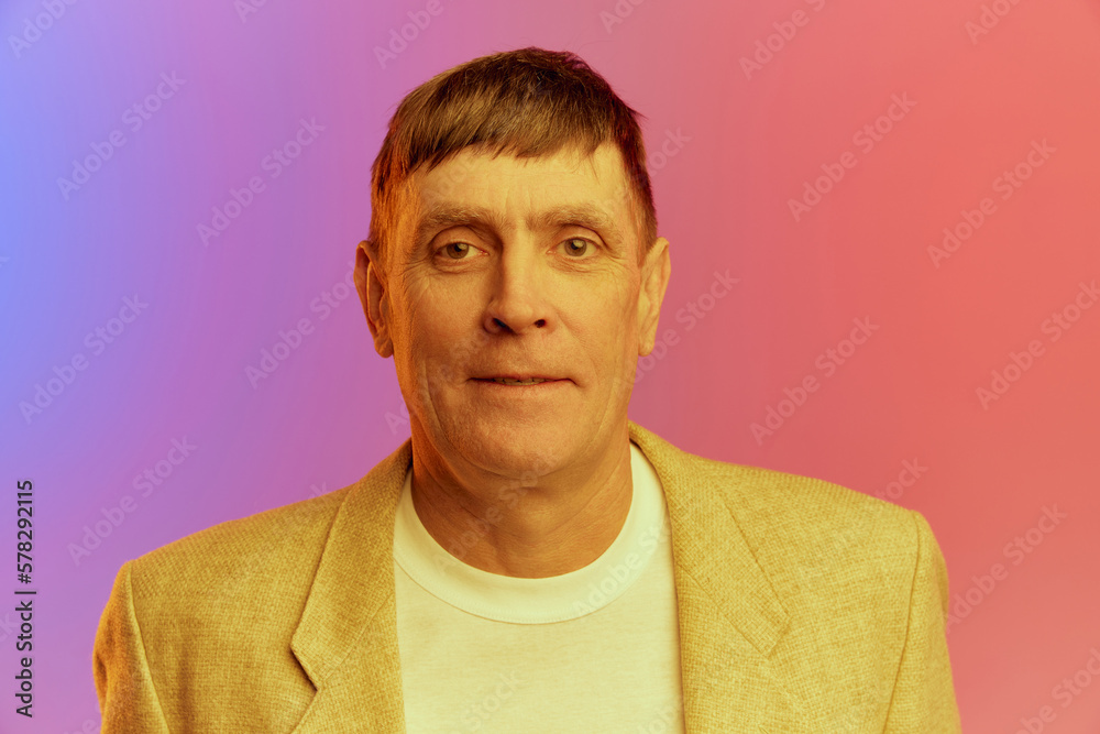 Portrait of mature, middle-aged man in jacket posing, looking at camera over gradient pink purple background in neon light. Concept of emotions, lifestyle, facial expression, fun. Ad