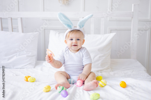 Wall Mural cute baby boy with bunny ears and colorful eggs on a white bed at home playing o
