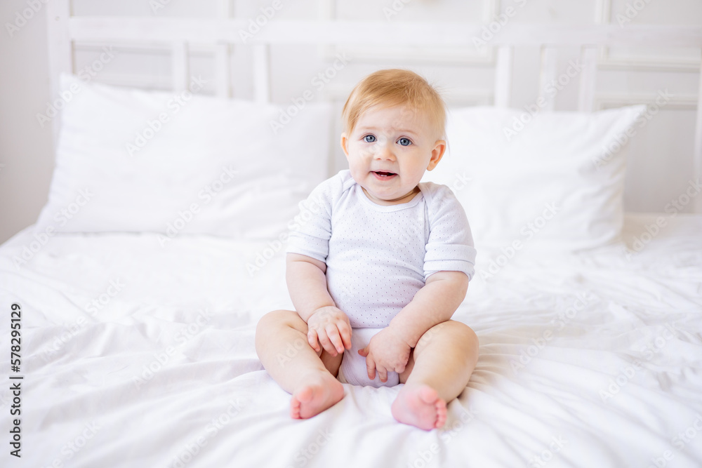 cute smiling baby boy is sitting on a white bed at home, a little blonde baby woke up in the morning, happy childhood and family