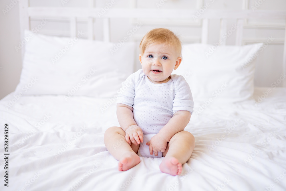 cute smiling baby boy is sitting on a white bed at home, a little blonde baby woke up in the morning, happy childhood and family cute smiling baby boy is sitting on a white bed at home, a little blonde baby woke up in the morning, happy childhood and family