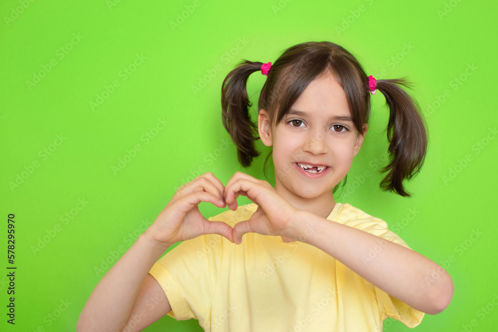 Close-up portrait of happy cute little toddler girl with missing baby tooth smile showing heart shape with hands on green background with copy space. Close-up portrait of happy cute little toddler girl with missing baby tooth smile showing heart shape with hands on green background with copy space.