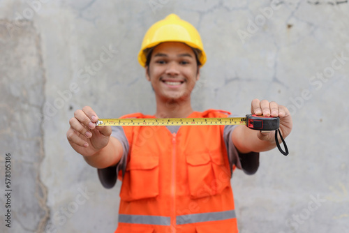 Excited contractor man in helmet showing measurement tool against wall background. 