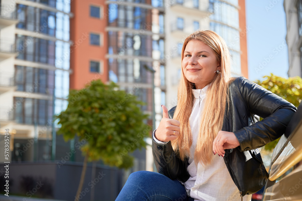 Portrait view of young pretty woman showing thumbs up, while sitting on bench at modern courtyard of city residential high-rise buildings.