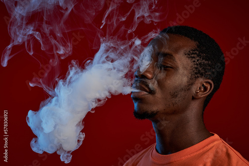 Young black man in orange t-shirt smoking e-cigarette in front of camera against red background and exhaling cloud of white smoke