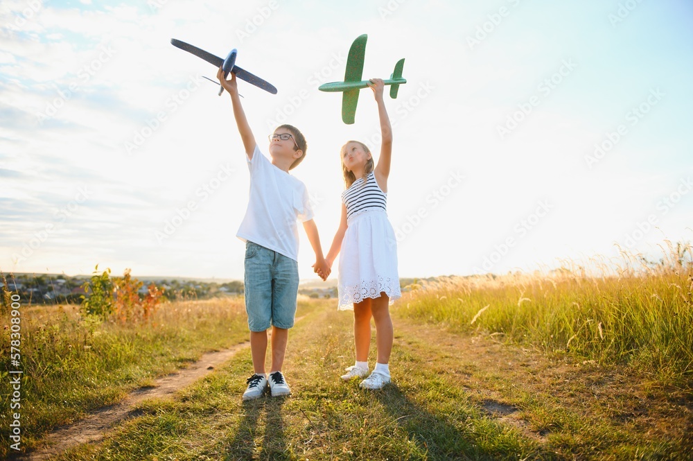 Obraz premium Running boy and girl holding two green and blue airplanes toy in the field during summer sunny day