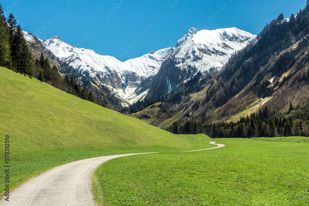 Beautiful landscape with snow capped mountains, green grass meadows and hiking trail in springtime. Trettachtal, Allgaeu, Bavaria, Germany. Beautiful landscape with snow capped mountains, green grass meadows and hiking trail in springtime. Trettachtal, Allgaeu, Bavaria, Germany.
