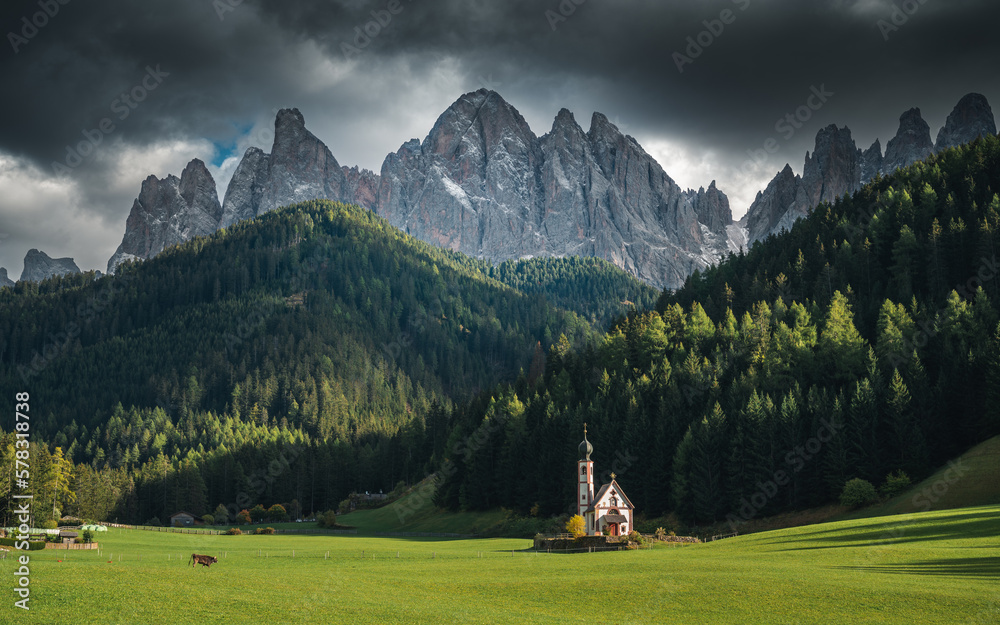 Tranquil scene from the chiesetta di San Giovanni (Funes), Dolomites, Italy. Green meadow with animals and snowy peaks with dark clouds
