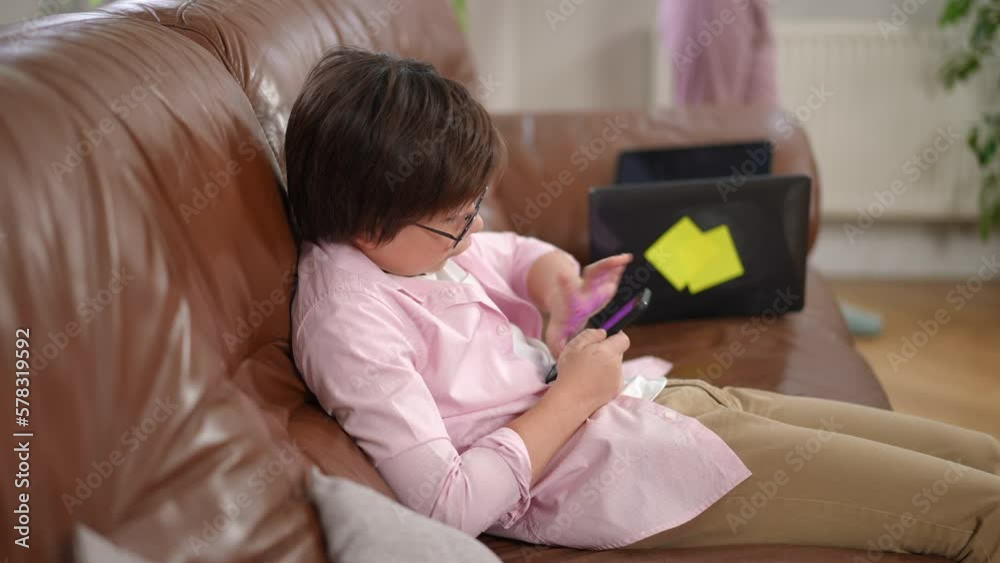 Side view carefree Caucasian boy in eyeglasses gaming in smartphone app sitting on couch in living room. Relaxed child enjoying leisure at home indoors Side view carefree Caucasian boy in eyeglasses gaming in smartphone app sitting on couch in living room. Relaxed child enjoying leisure at home indoors