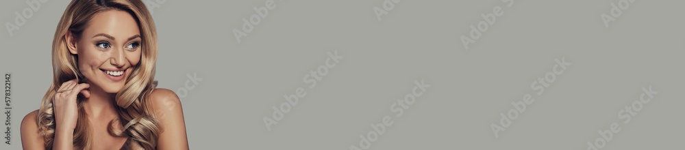 Beautiful young woman with long and waving blond hair smiling against grey background Beautiful young woman with long and waving blond hair smiling against grey background