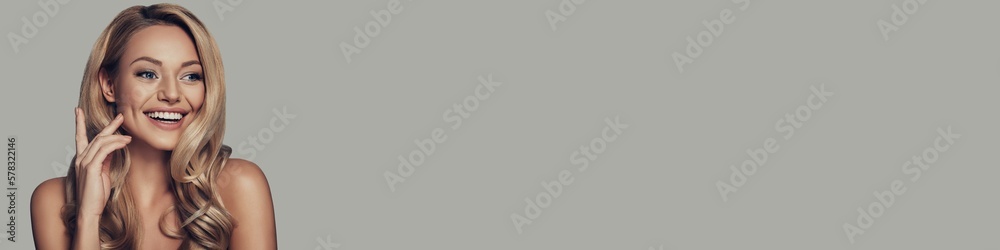Attractive young woman with long and waving blonde hair smiling against grey background Attractive young woman with long and waving blonde hair smiling against grey background