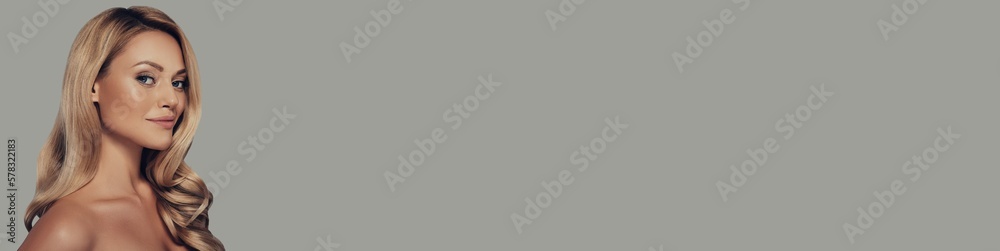 Attractive young woman with long and waving blonde hair looking at camera against grey background