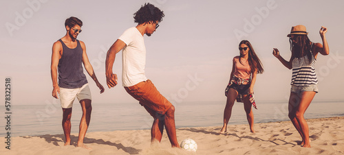 Group of cheerful young people playing with soccer ball on the beach with a sea in the background