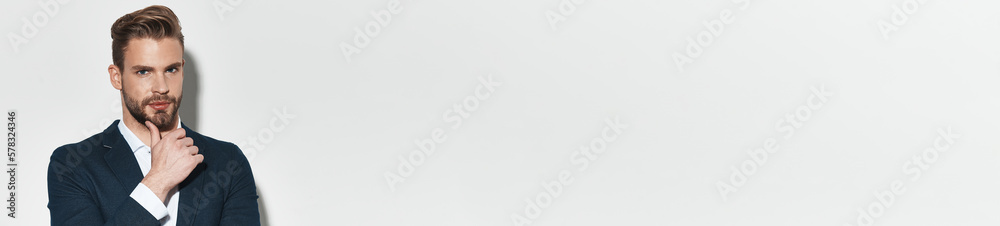 Handsome young man in suit holding hand on chin while standing against grey background Handsome young man in suit holding hand on chin while standing against grey background