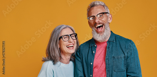Fotografi Beautiful senior couple in eyeglasses laughing while standing against colored ba