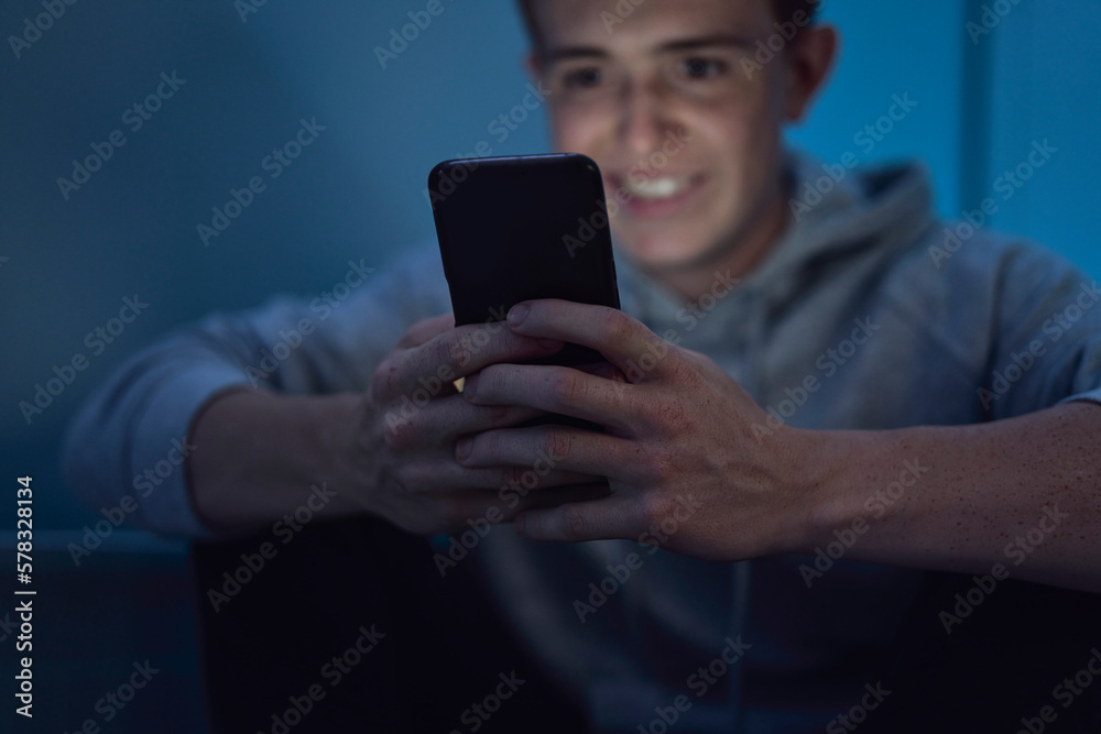 Cheerful caucasian teenage boy using mobile phone while sitting at night in his room