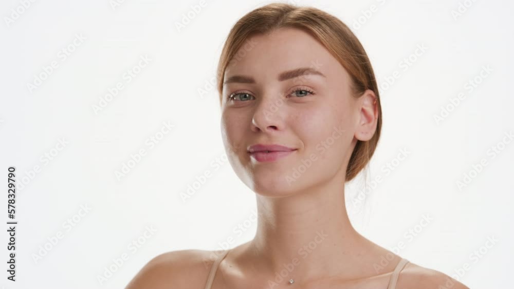 Face care cosmetics commercial | Close-up shot of cute young slim brown-haired Caucasian woman strokes her cheek looking at the camera and smiling for it on white background Face care cosmetics commercial | Close-up shot of cute young slim brown-haired Caucasian woman strokes her cheek looking at the camera and smiling for it on white background