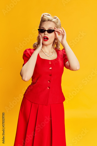 Beautiful young girl with stylish hairstyle in red suit and trendy sunglasses posing with shocked face against yellow studio background. Concept of retro fashion  beauty  50s  60s. Pin-up style