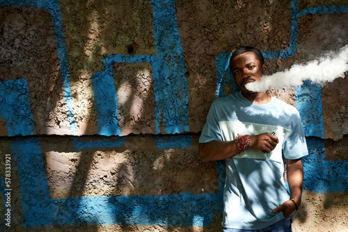 African American guy in blue t-shirt smoking vape pen on sunny day while standing against wall of building in shadow and looking at camera © Seventyfour