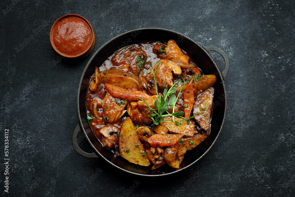 Fried potatoes in sauce, with mushrooms and vegetables, on dark concrete