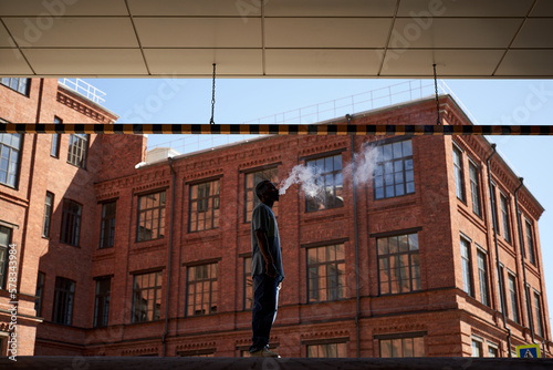 Side view of smoking young black man in casualwear standing against red brick building and releasing cloud of vapor out of mouth