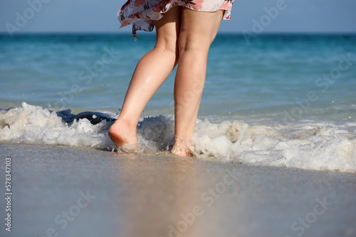 Barefoot woman in short dress walking by the sand in sea waves. Female legs in water splashes, beach vacation